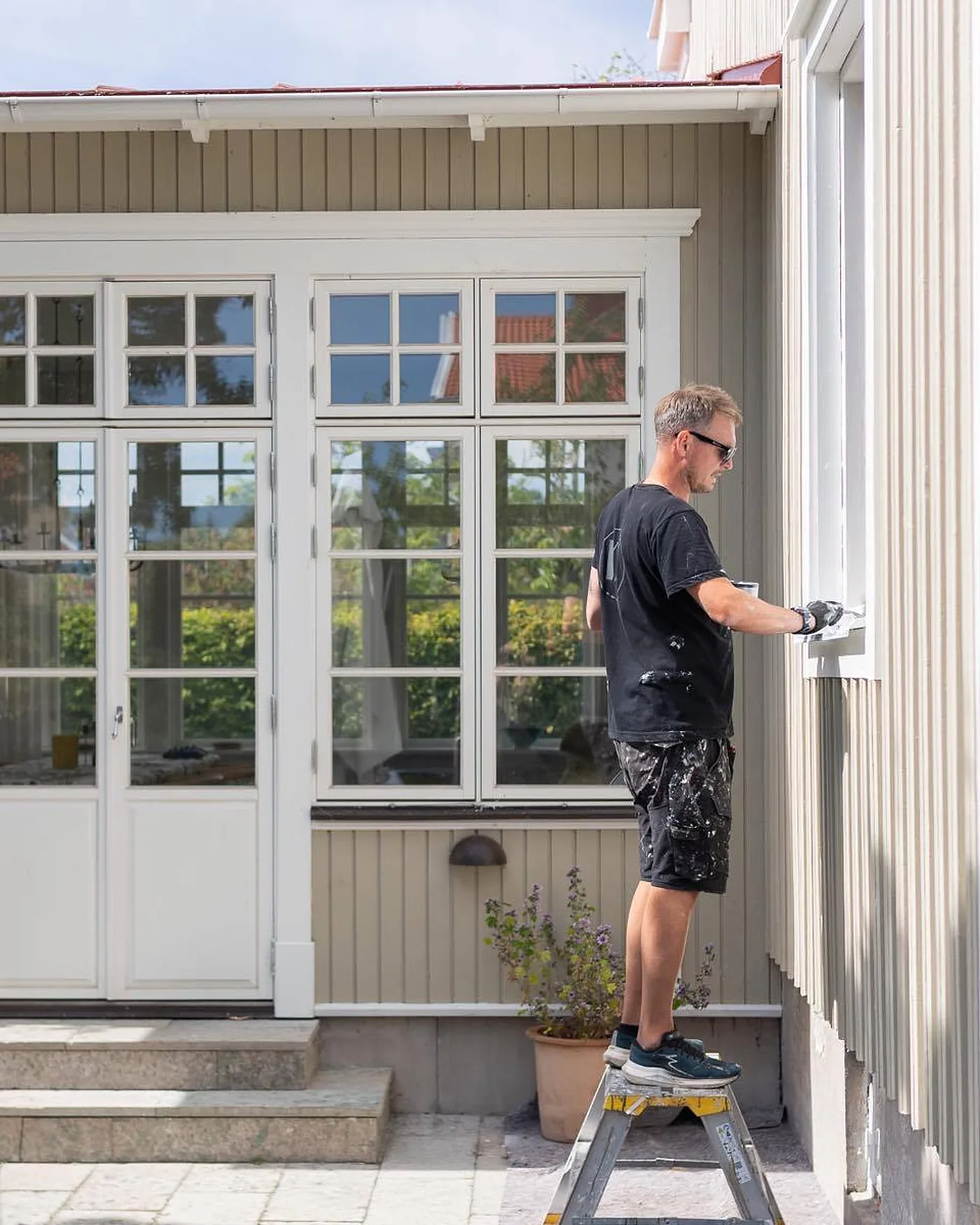 Målare från KA Måleriservice på en pall som målar fönsterkarmen på en ljusgrå trävilla, med spröjsade glasdörrar bredvid
